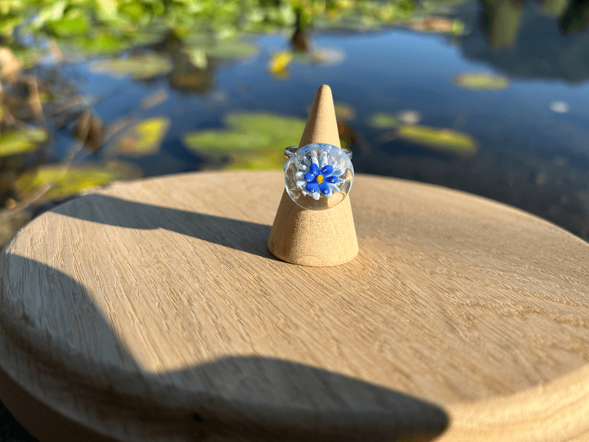 Bague cabochon en verre ornée d'une fleur bleue posée sur un présentoir conique en bois.