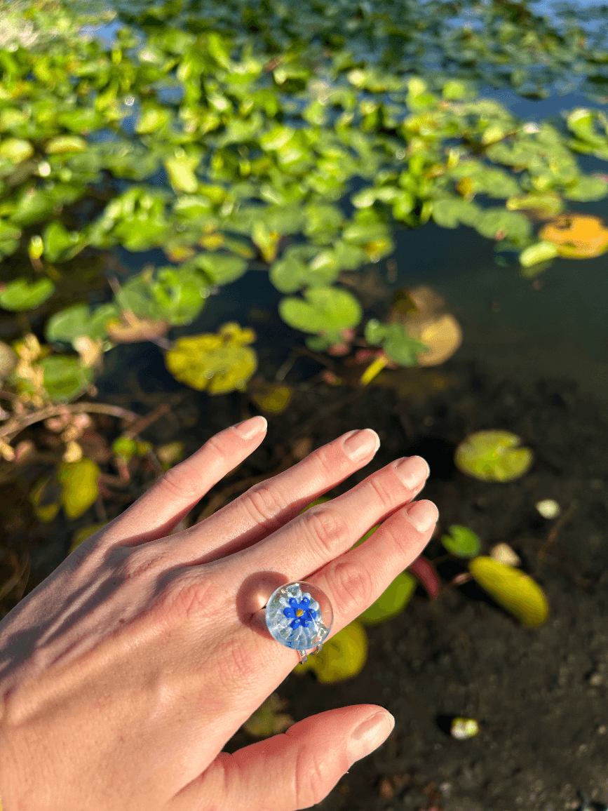Bague en verre transparent portée à la main, révélant une fleur bleue et jaune, devant un étang.