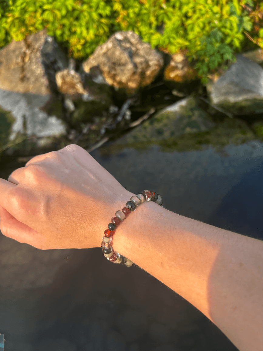 Bracelet en perles de verre ambrées, taupe et sombres porté au poignet près d'un point d'eau