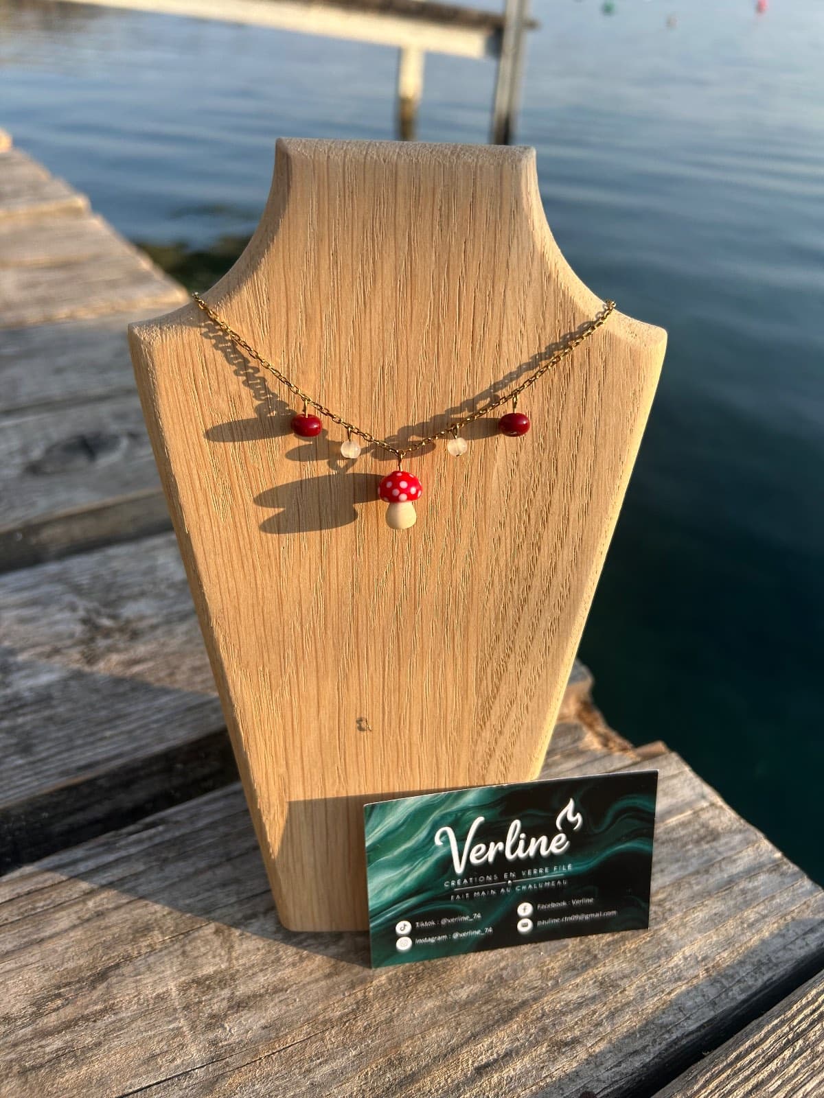 Collier à chaîne dorée et pendentif champignon rouge à pois blancs exposé sur un buste en bois face à un lac.
