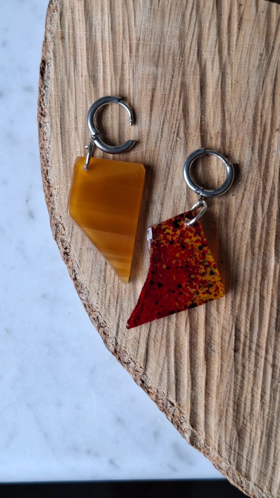 Boucles d'oreilles pendantes en verre jaune et rouge moucheté, présentées à plat sur une rondelle de bois.