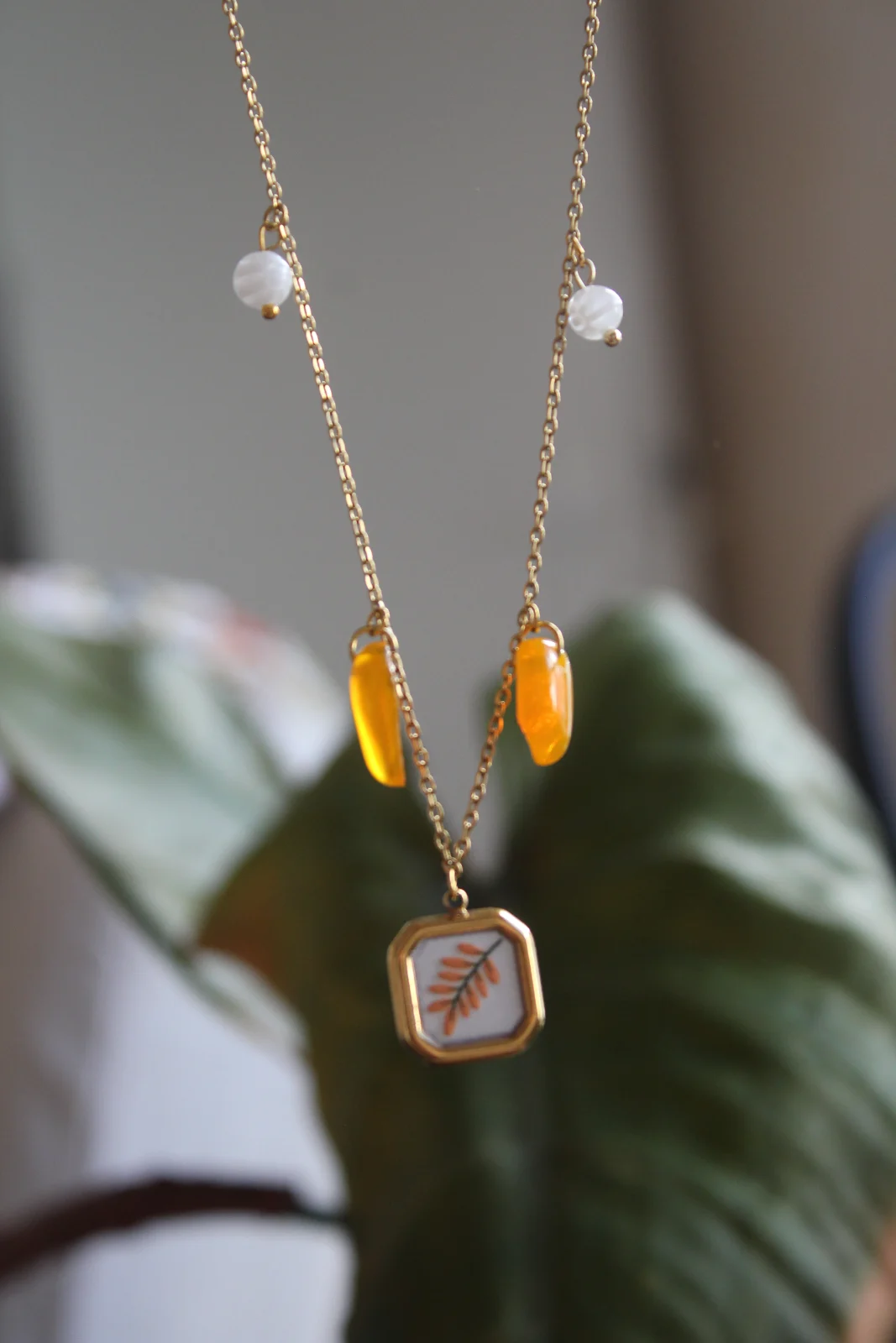Détail du collier doré suspendu avec pampilles orange, perles blanches et pendentif feuillu.