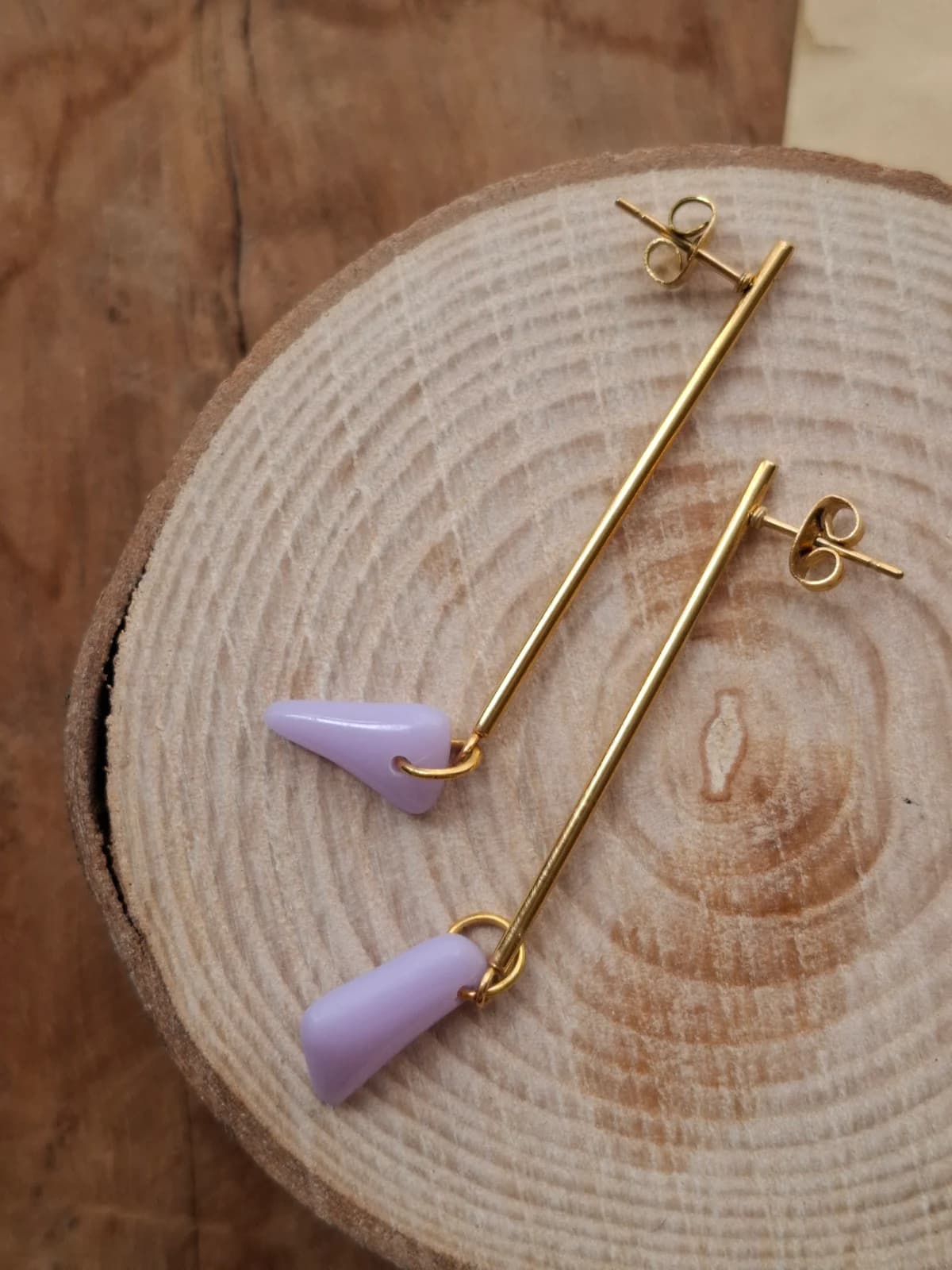Paire de boucles d'oreilles pendantes à tiges dorées et gouttes violettes posées sur un rondin de bois.