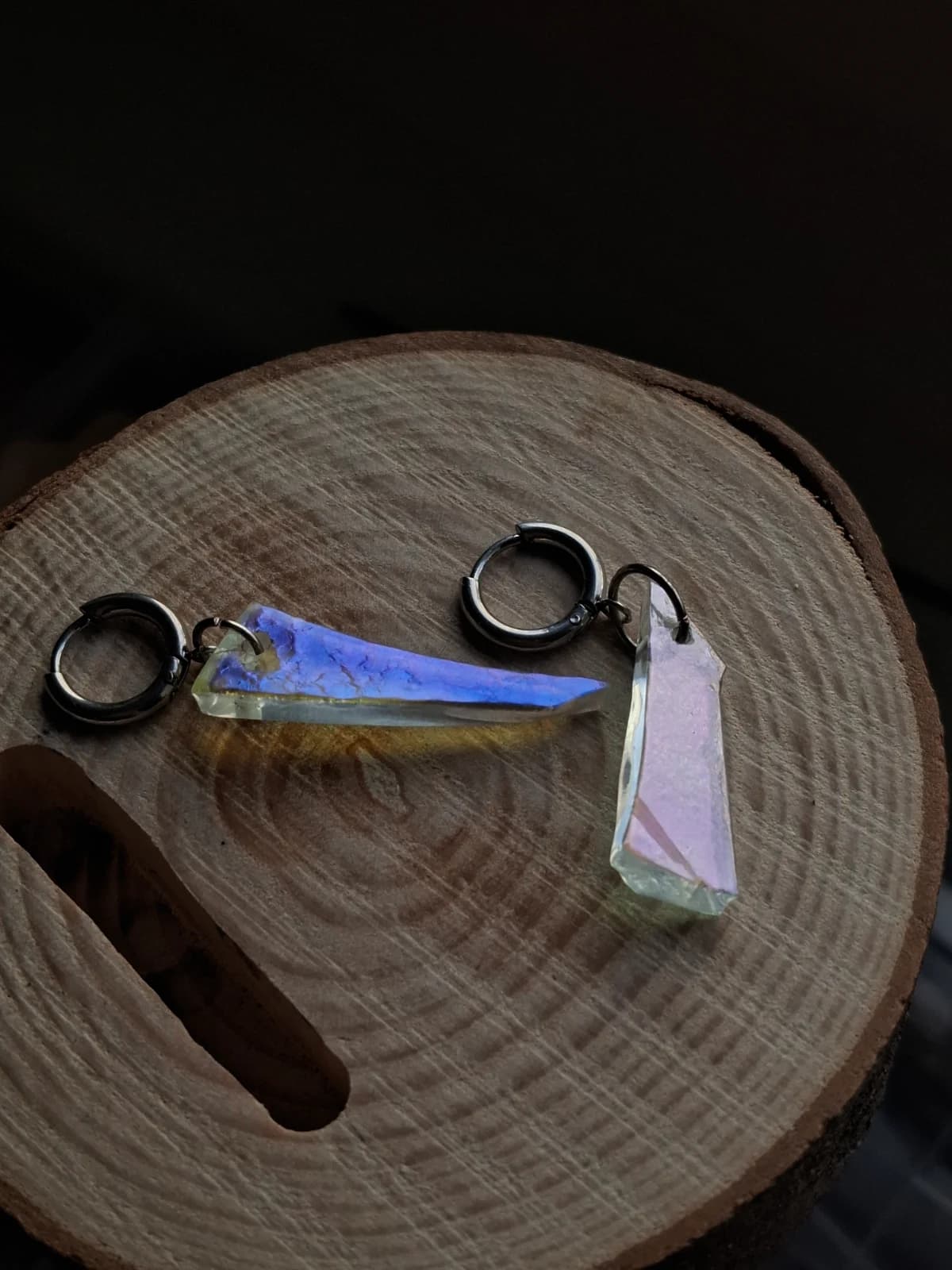 Boucles d'oreilles asymétriques en verre aux reflets bleutés et mauves éclairées dans la pénombre.