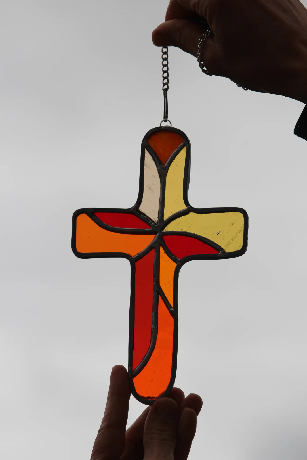 Croix en vitrail aux teintes orangées, rouges et jaunes tenue à la main en contre-jour