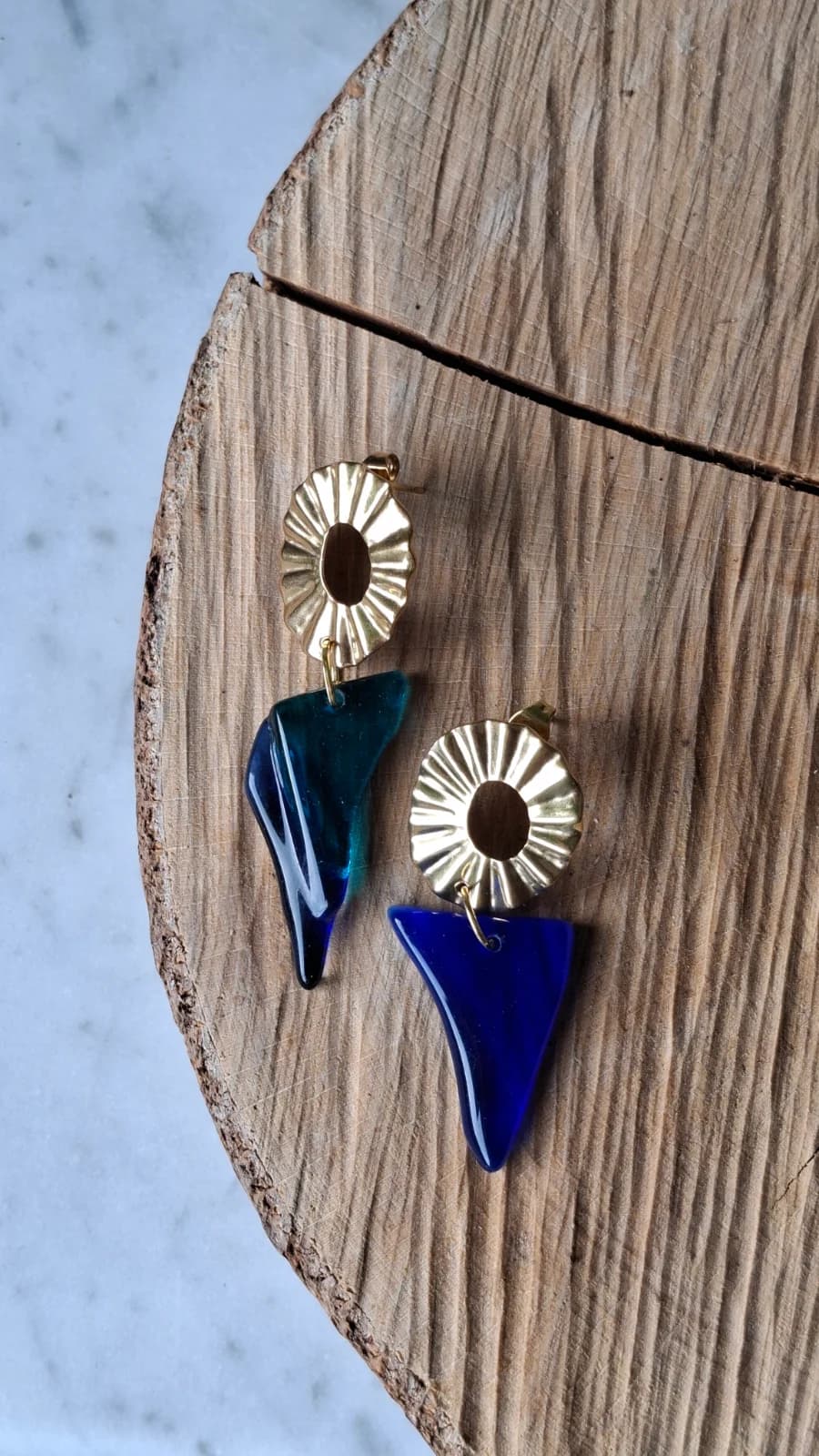 Boucles d'oreilles dorées texturées avec pendentifs asymétriques en verre bleu posées sur du bois.