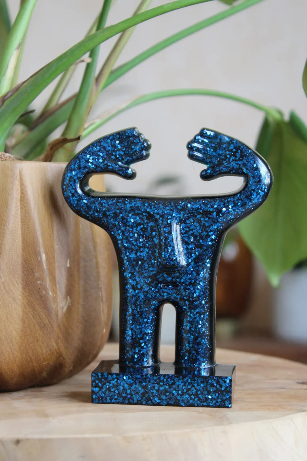 Statuette stylisée bleue à paillettes scintillantes aux bras levés sur socle en bois.