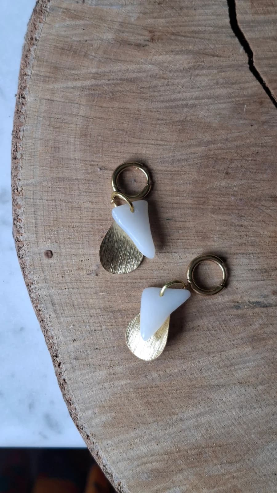 Boucles d'oreilles dorées en verre blanc et pampilles texturées posées à plat sur une tranche de bois.