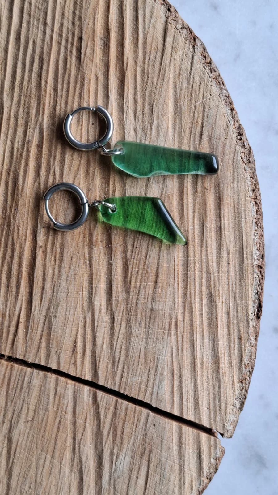 Boucles d'oreilles avec pendentifs en verre vert aux formes asymétriques posées sur un rondin de bois.