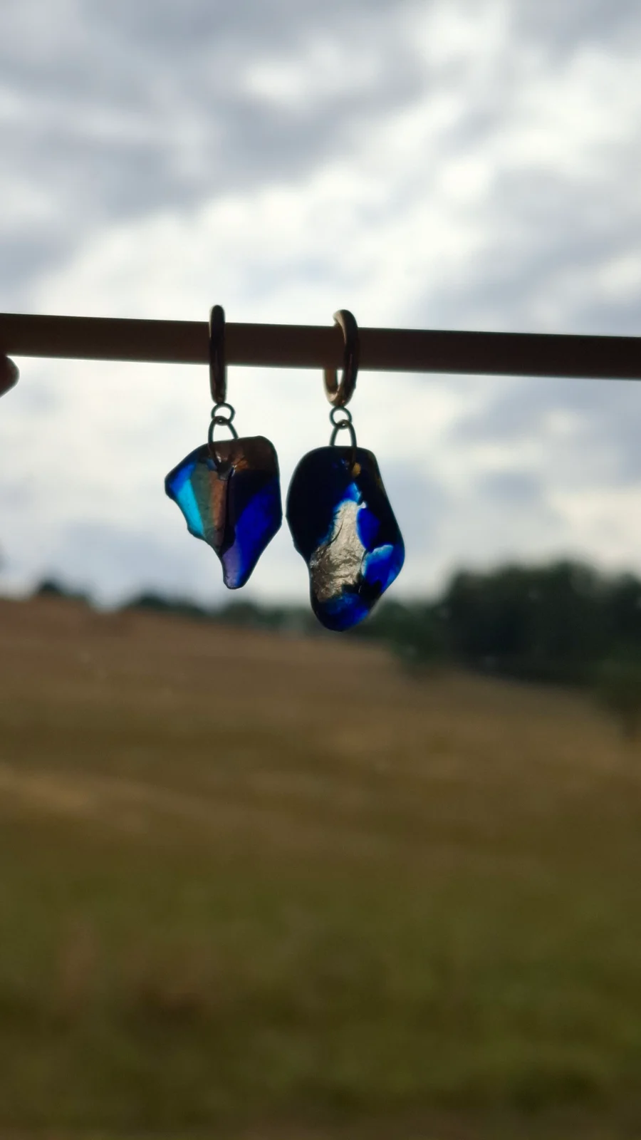 Boucles d'oreilles en verre bleu et transparent rétroéclairées devant un paysage naturel.