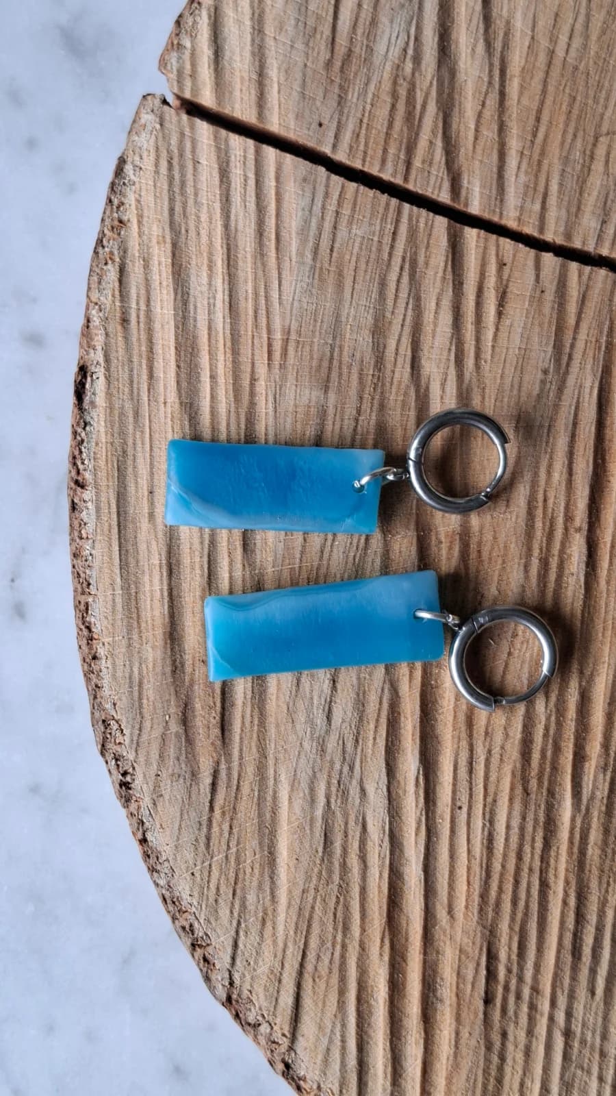 Paire de boucles d'oreilles pendantes en verre bleu et anneaux argentés posée à plat sur une tranche de bois.