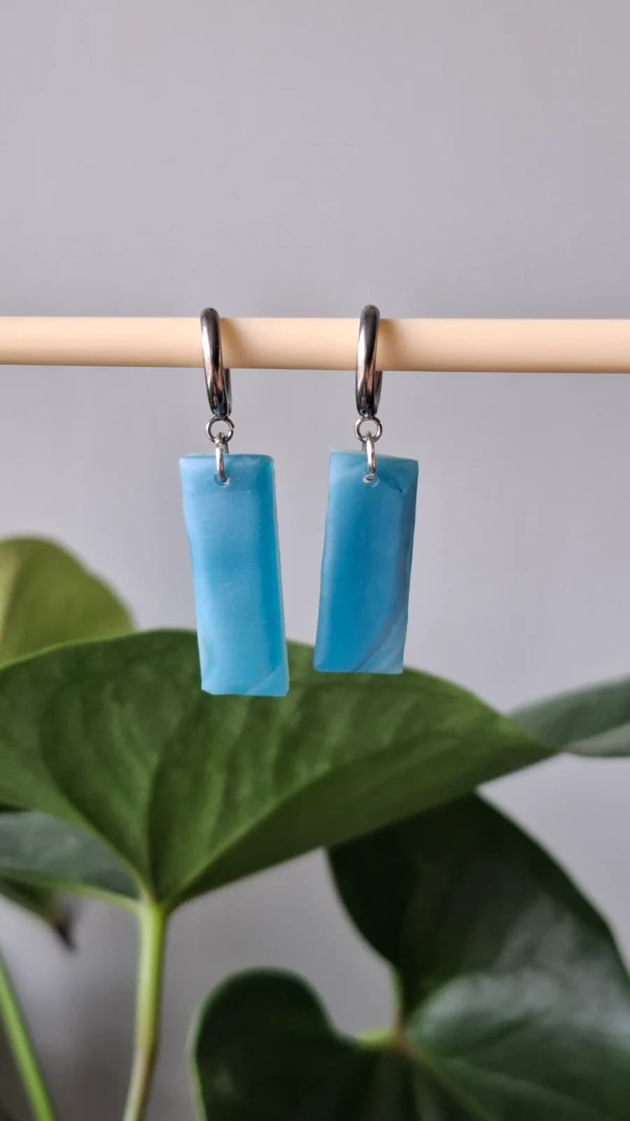 Boucles d'oreilles rectangulaires en verre bleu montées sur dormeuses, suspendues devant un feuillage vert.