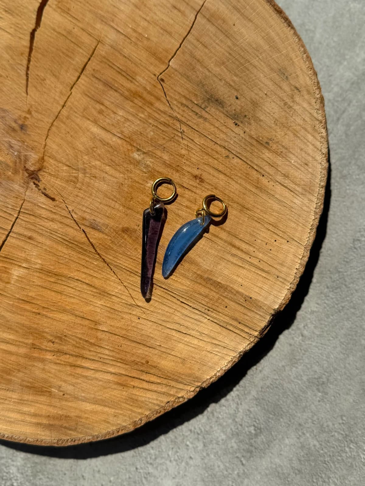 Boucles d'oreilles pendantes en verre violet et bleu avec attaches dorées posées sur un rondin de bois