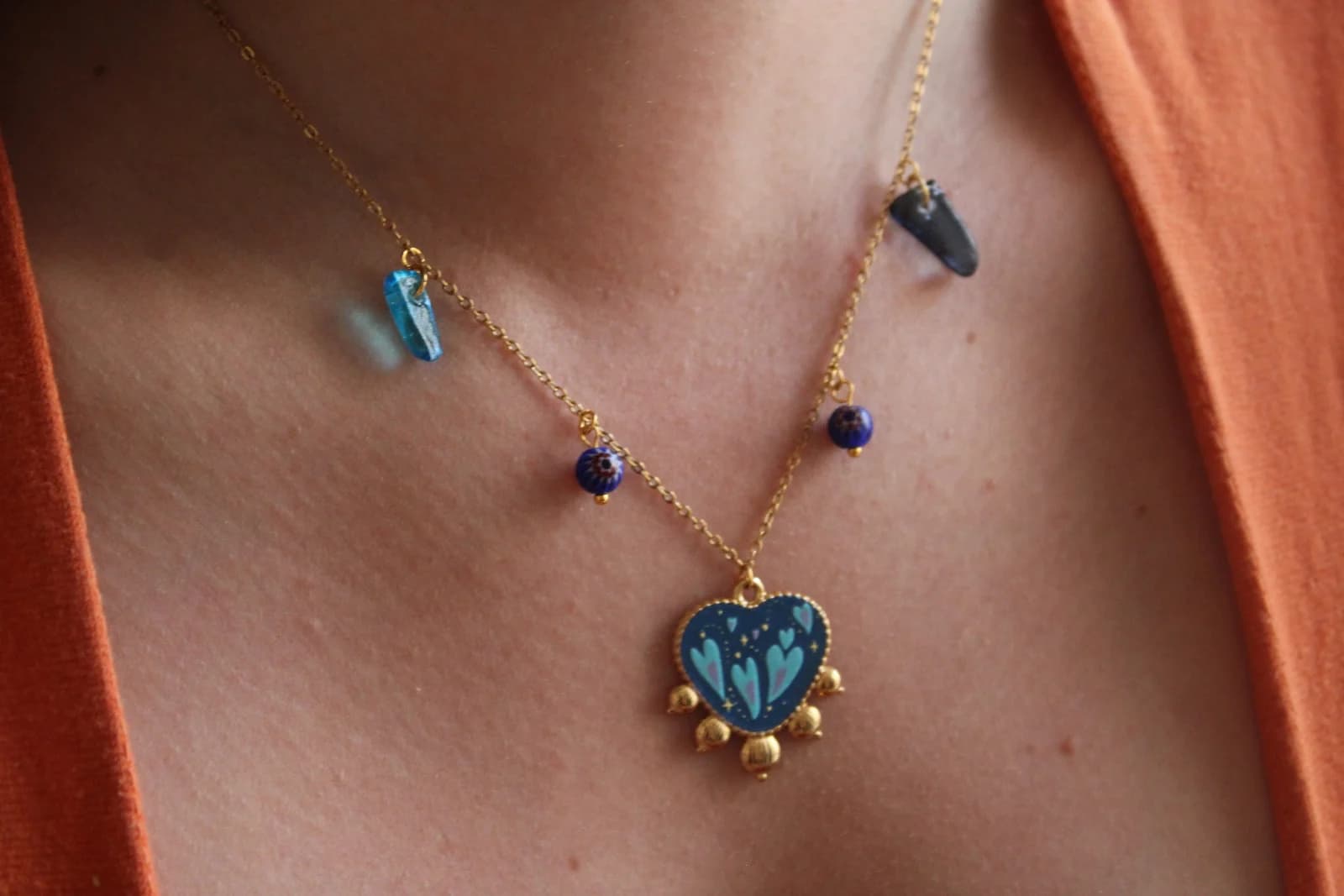Collier en chaîne dorée avec pendentif cœur bleu et pampilles porté sur un buste.