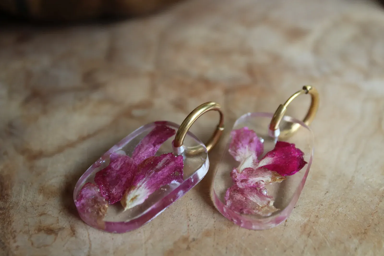 Boucles d'oreilles pendantes rectangulaires en résine transparente et pétales roses, posées sur du bois.