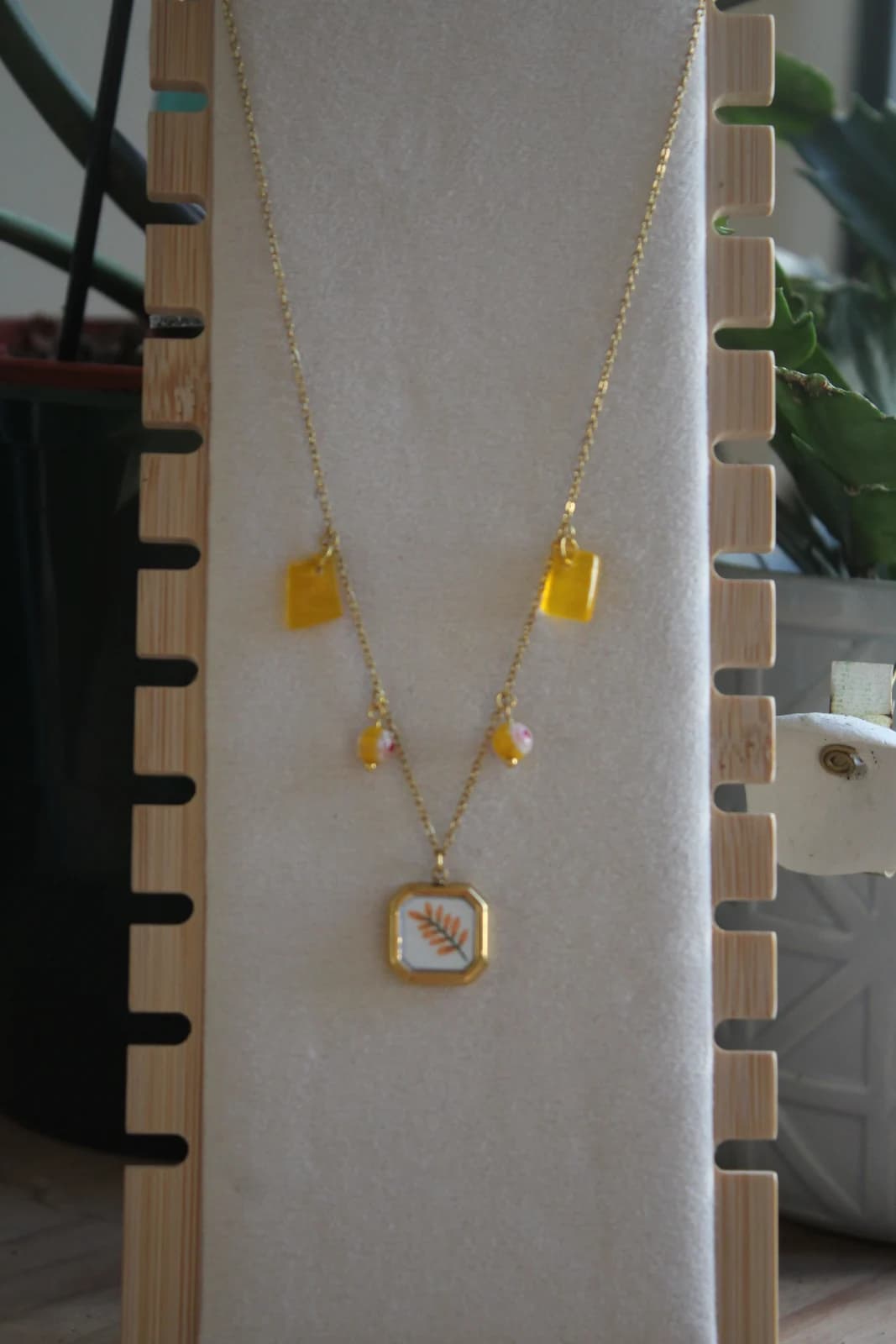 Collier doré avec pendentif carré à motif végétal, perles et pampilles jaunes sur présentoir.