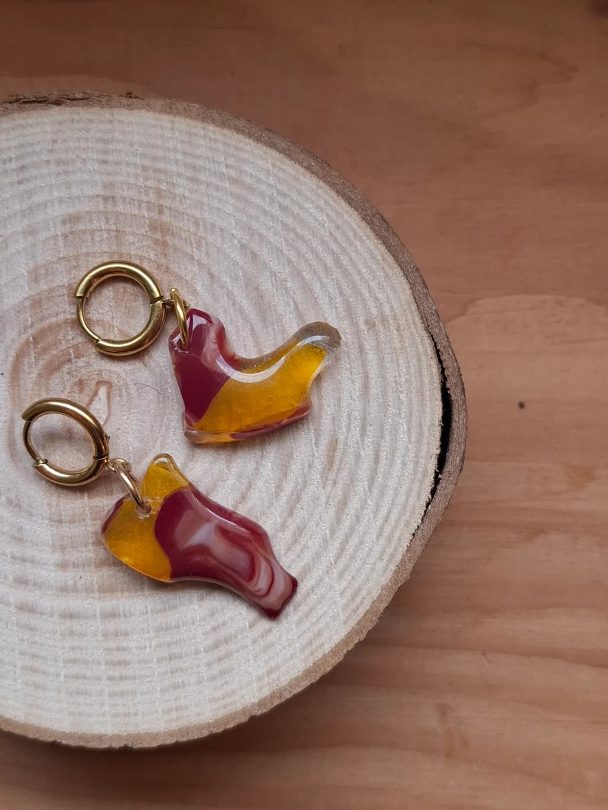 Boucles d'oreilles pendantes asymétriques en verre jaune et rouge posées sur une rondelle de bois