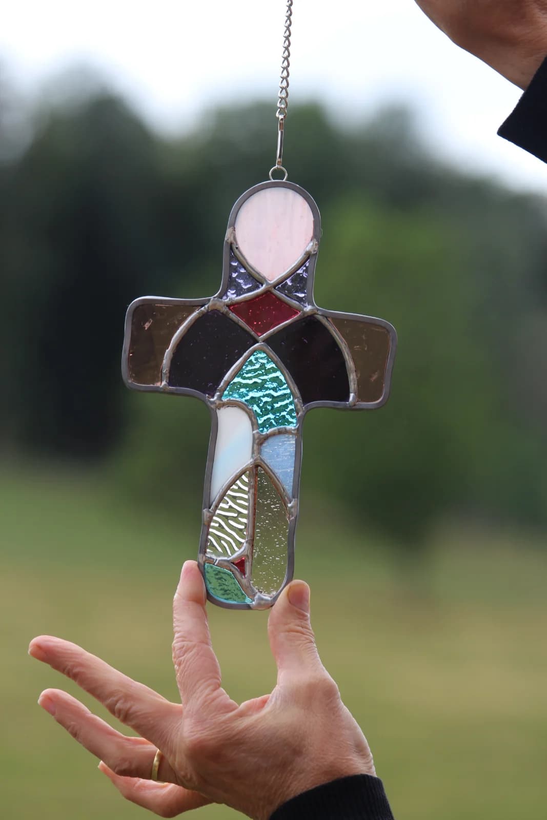 Croix en vitrail multicolore et texturé suspendue par une chaînette devant un fond naturel.