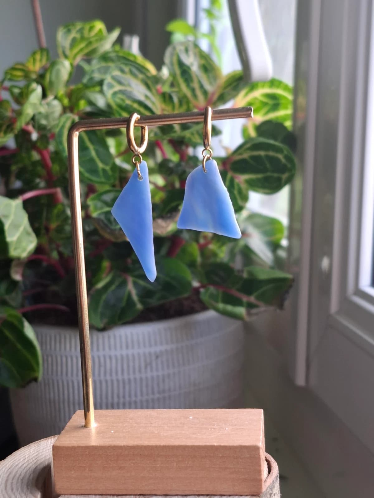 Boucles d'oreilles pendantes en verre bleu asymétrique suspendues à un présentoir doré devant une plante.