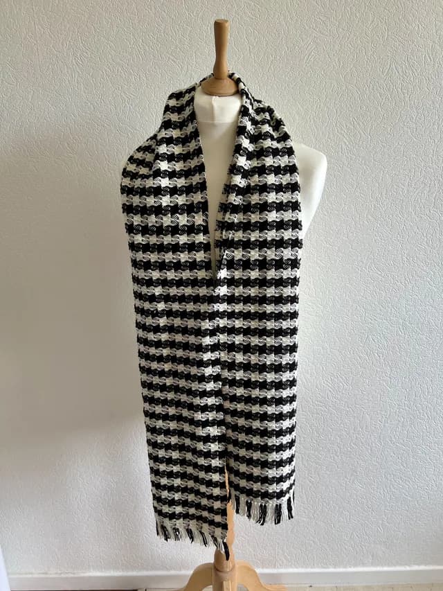 Écharpe tissée à motifs géométriques noirs et blancs avec franges, drapée sur un buste de couture.