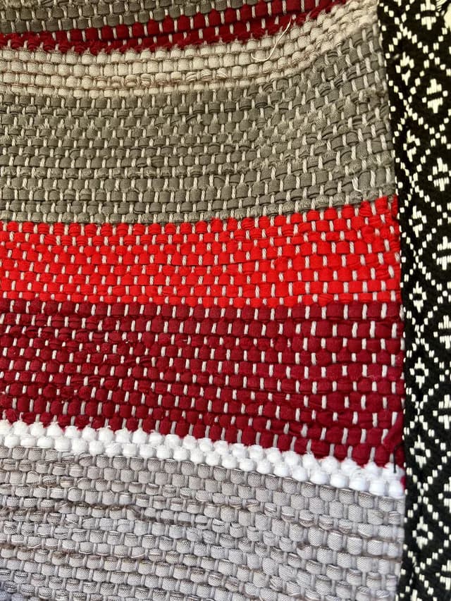 Détail du tissage artisanal aux rayures rouges, bordeaux et grises avec galon noir et blanc.