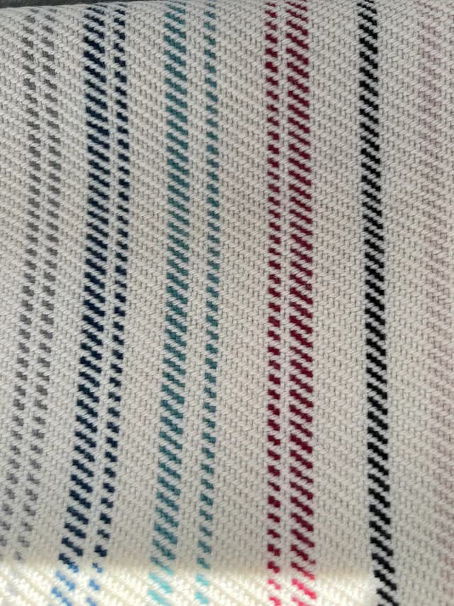 Gros plan sur le tissage écru et les rayures verticales grises, bleues, turquoise, rouges et noires.