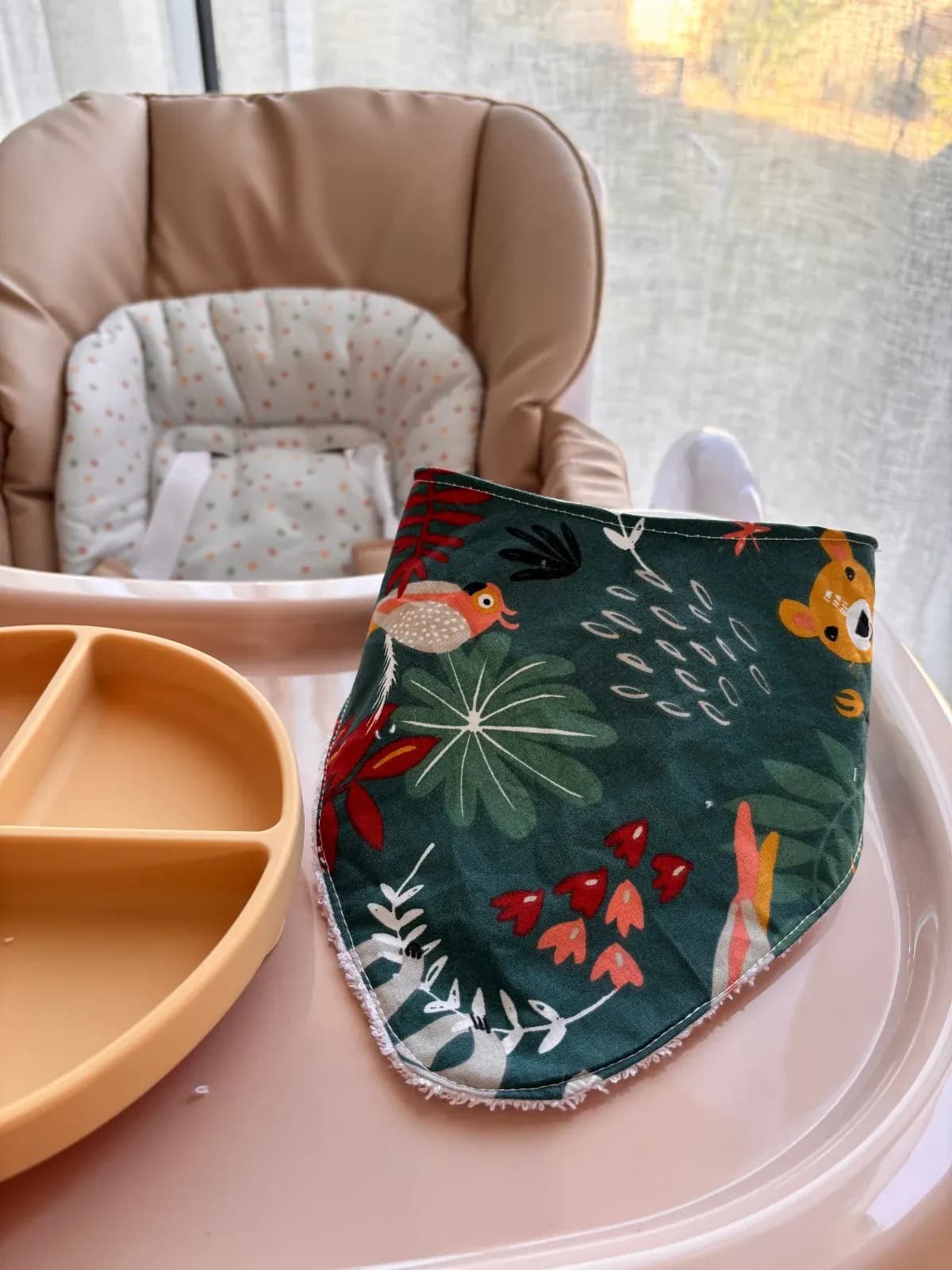 Bavoir bandana vert déplié à motifs de jungle et animaux à côté d'une assiette sur une tablette pour bébé.