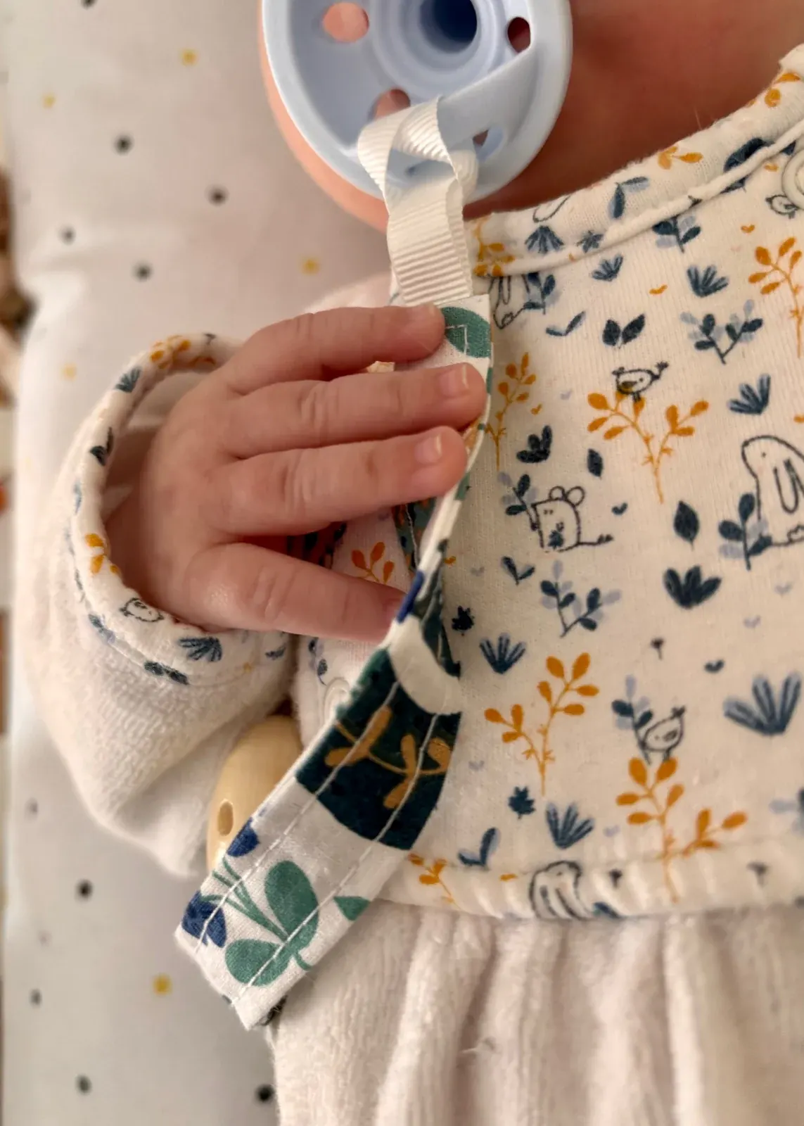 Attache-tétine en tissu à motifs bleus et verts avec clip en bois clair, posée sur un tissu doux écru.