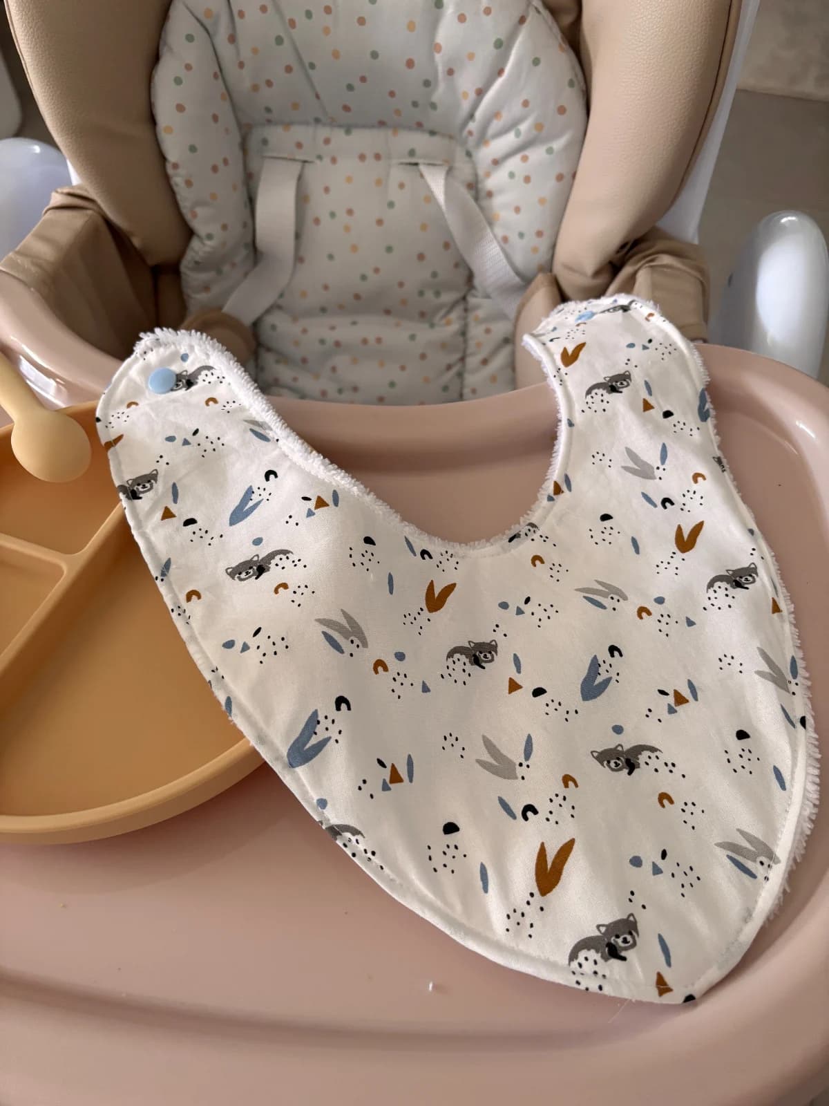 Bavoir bandana étalé montrant ses motifs de ratons laveurs, son bouton pression bleu clair et sa coupe triangulaire.