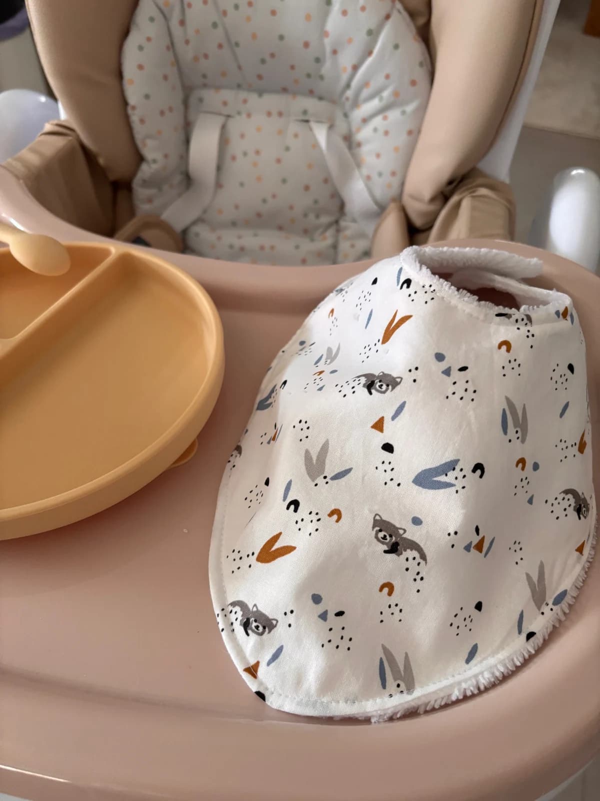 Bavoir bandana blanc à motifs animaux des bois et bordure éponge, posé sur le plateau d'une chaise haute.