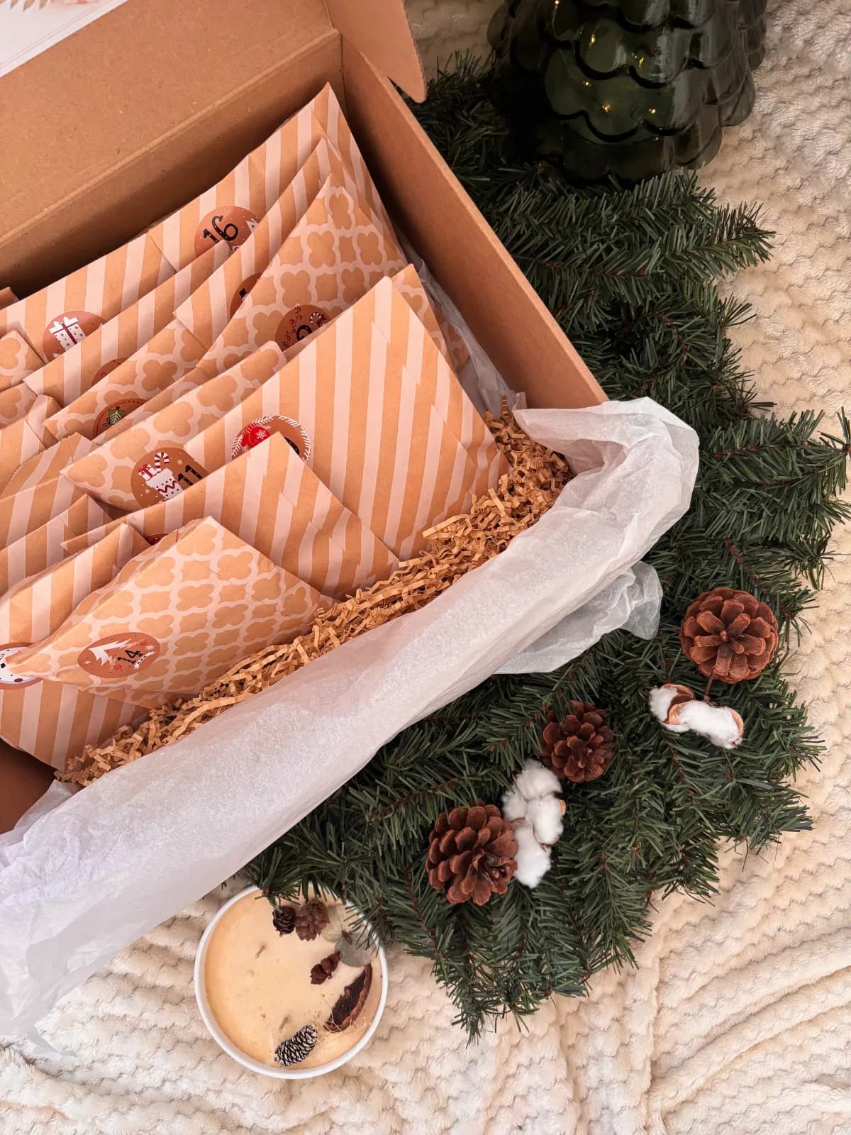 Boîte kraft ouverte garnie de pochettes numérotées, papier de soie blanc et décoration végétale d'hiver