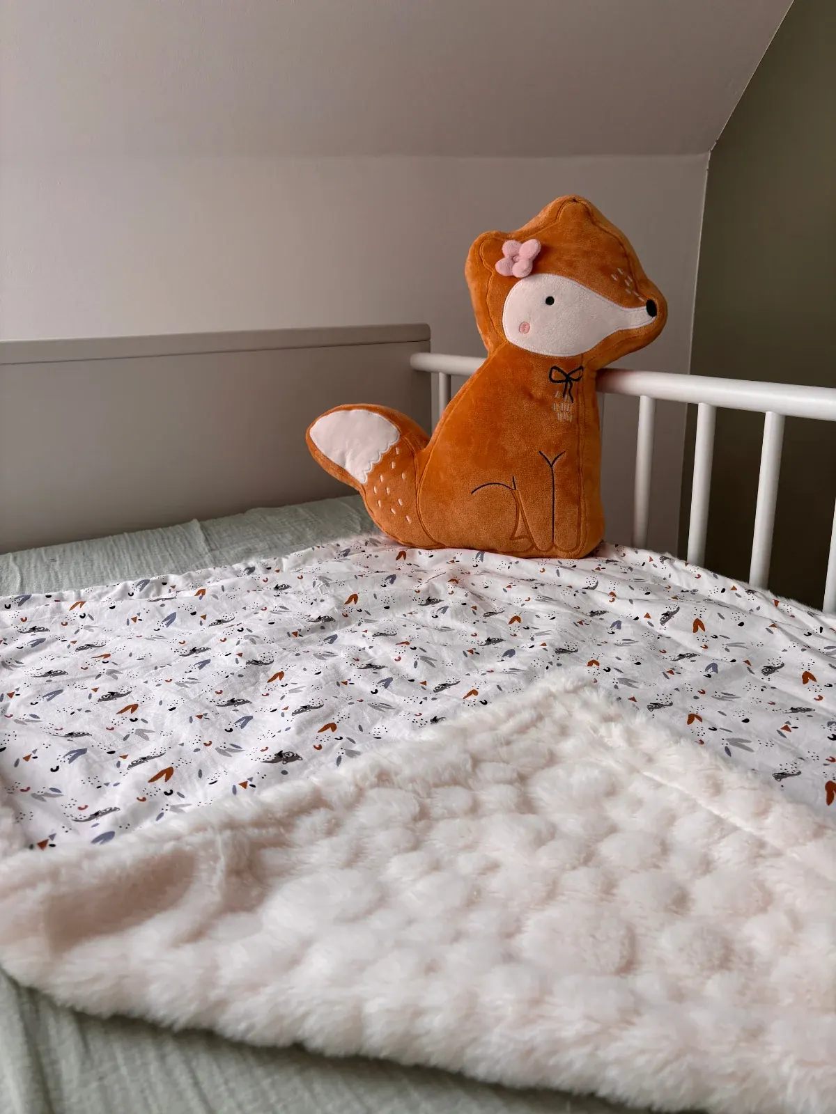 Couverture bébé dépliée sur un lit d'enfant à côté d'un coussin en forme de renard roux.