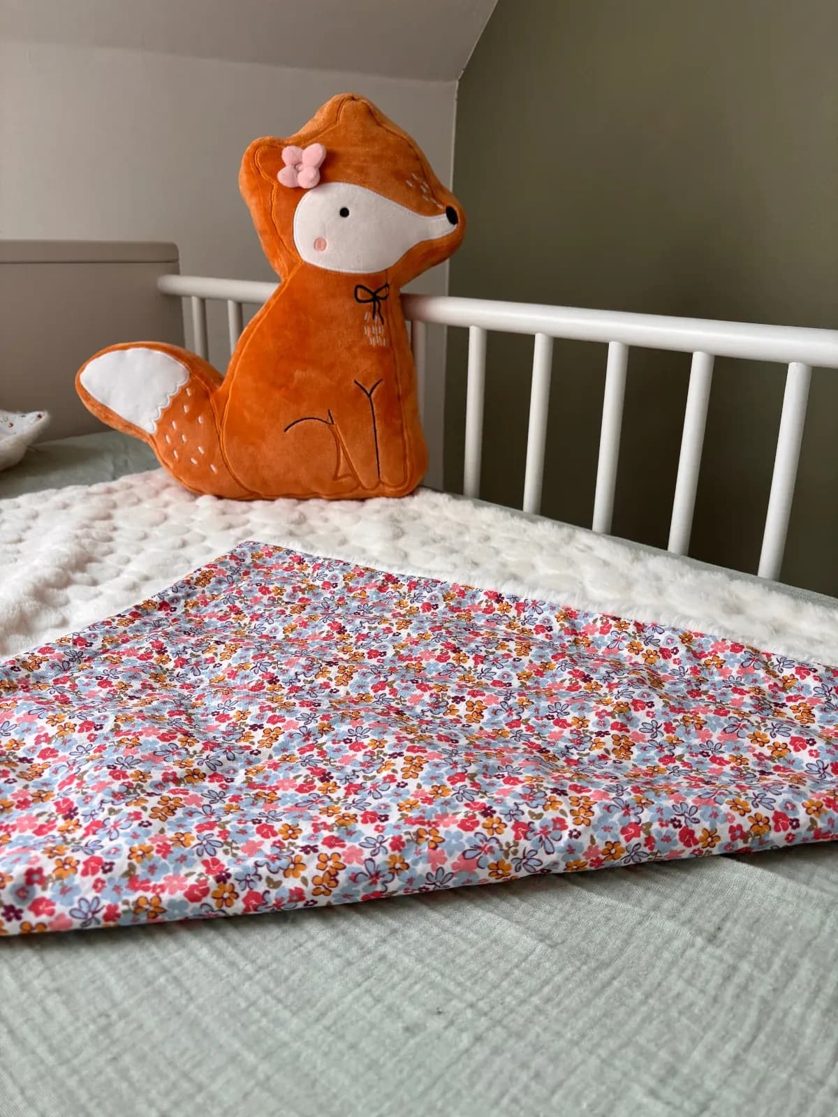 Couverture bébé à motif floral et polaire blanche texturée sur un lit d'enfant à côté d'un coussin renard.