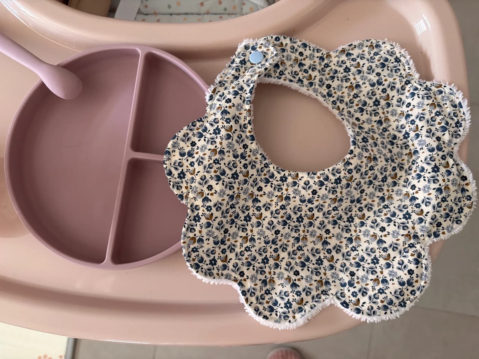 Bavoir bébé en forme de fleur à motif fleuri bleu, posé à côté d'une assiette rose