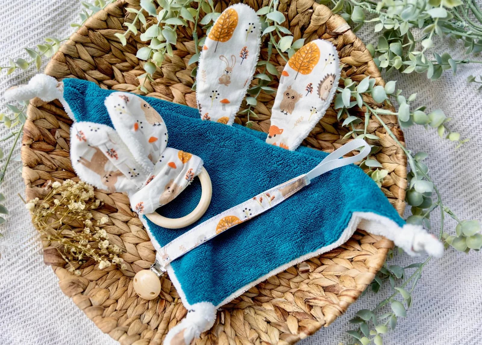Doudou plat bleu canard à oreilles de lapin, anneau hochet et attache-tétine aux motifs de la forêt dans une corbeille