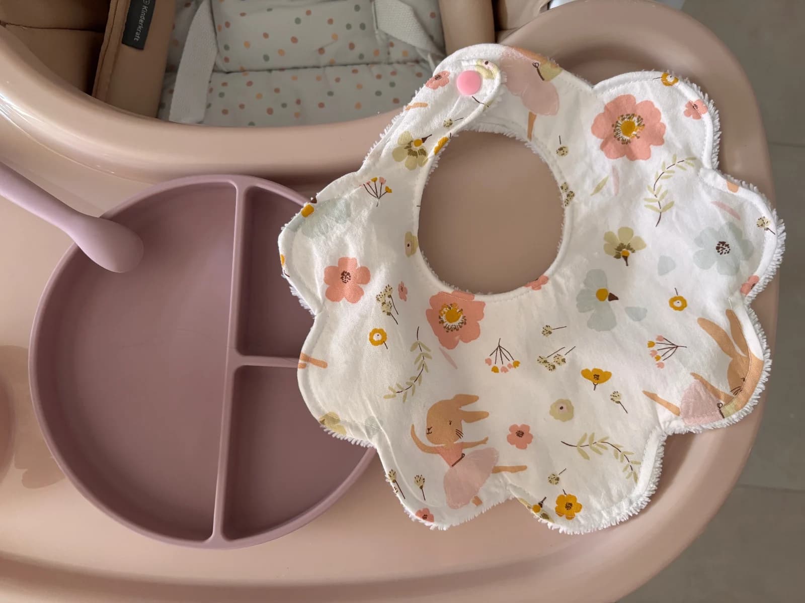 Bavoir blanc en forme de fleur aux motifs floraux et lapins, posé près d'une assiette à compartiments mauve.