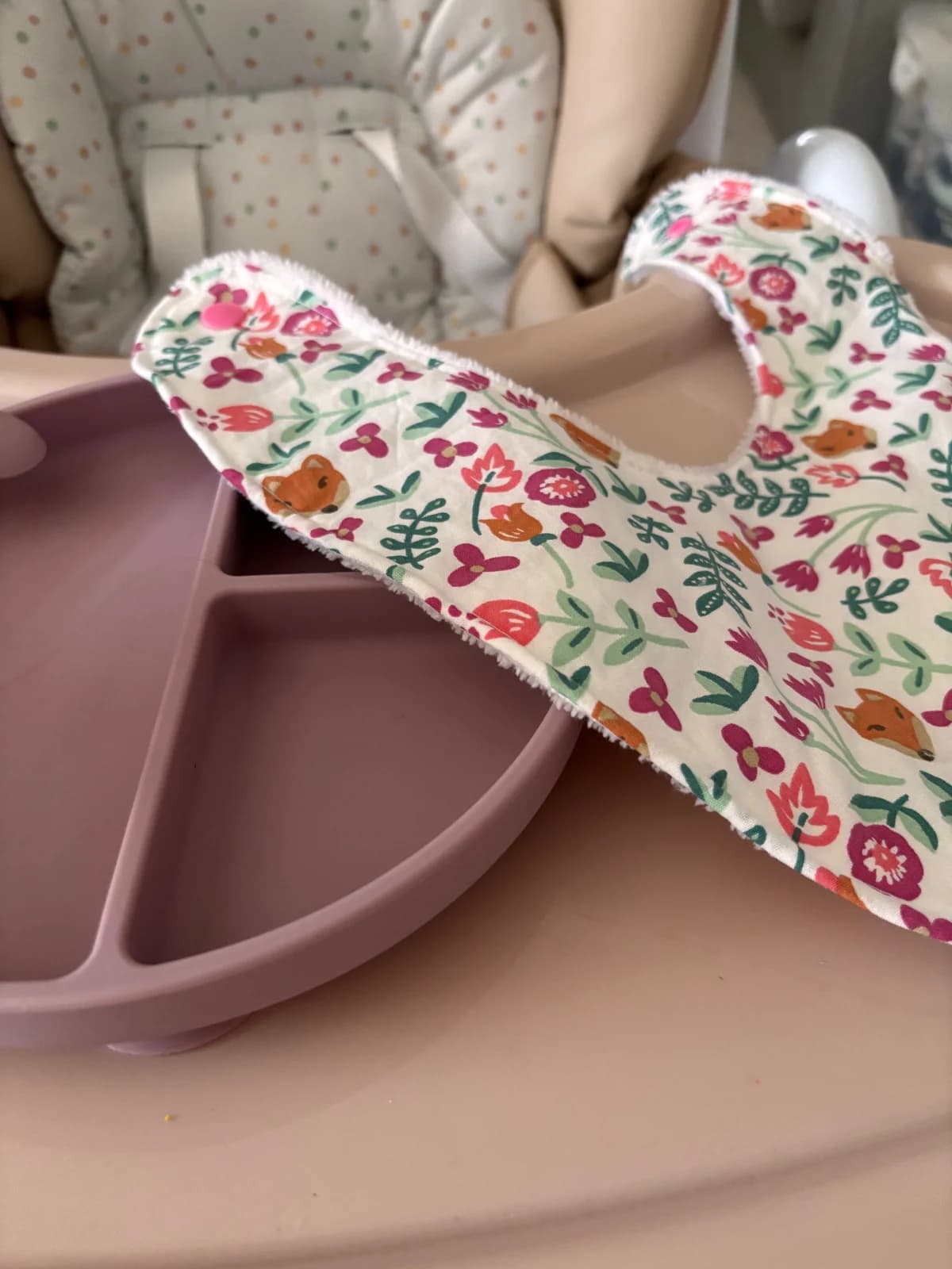 Bavoir bandana déplié à motifs de renardeaux et fleurs avec bouton-pression rose, à côté d'une assiette.