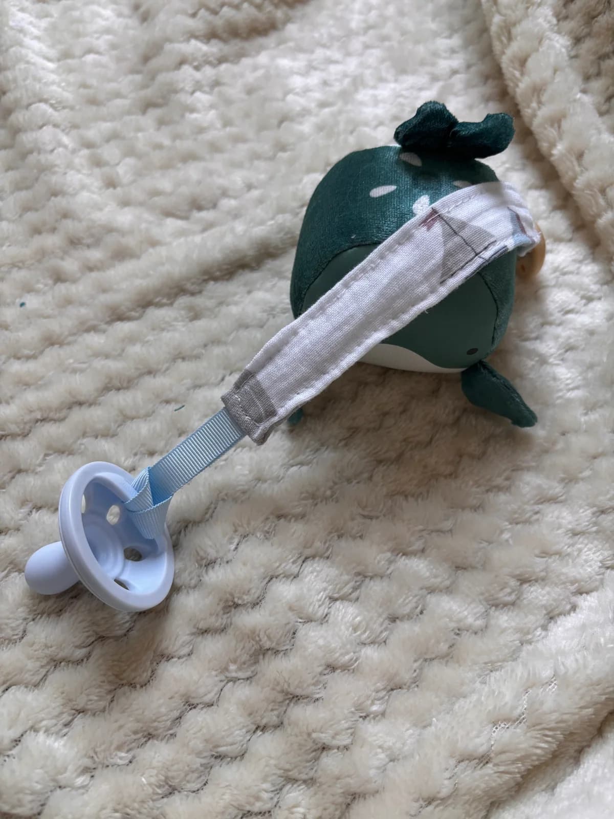 Vue de dessus de l'attache-tétine en tissu clair fixée à une tétine par un ruban bleu sur un plaid blanc.