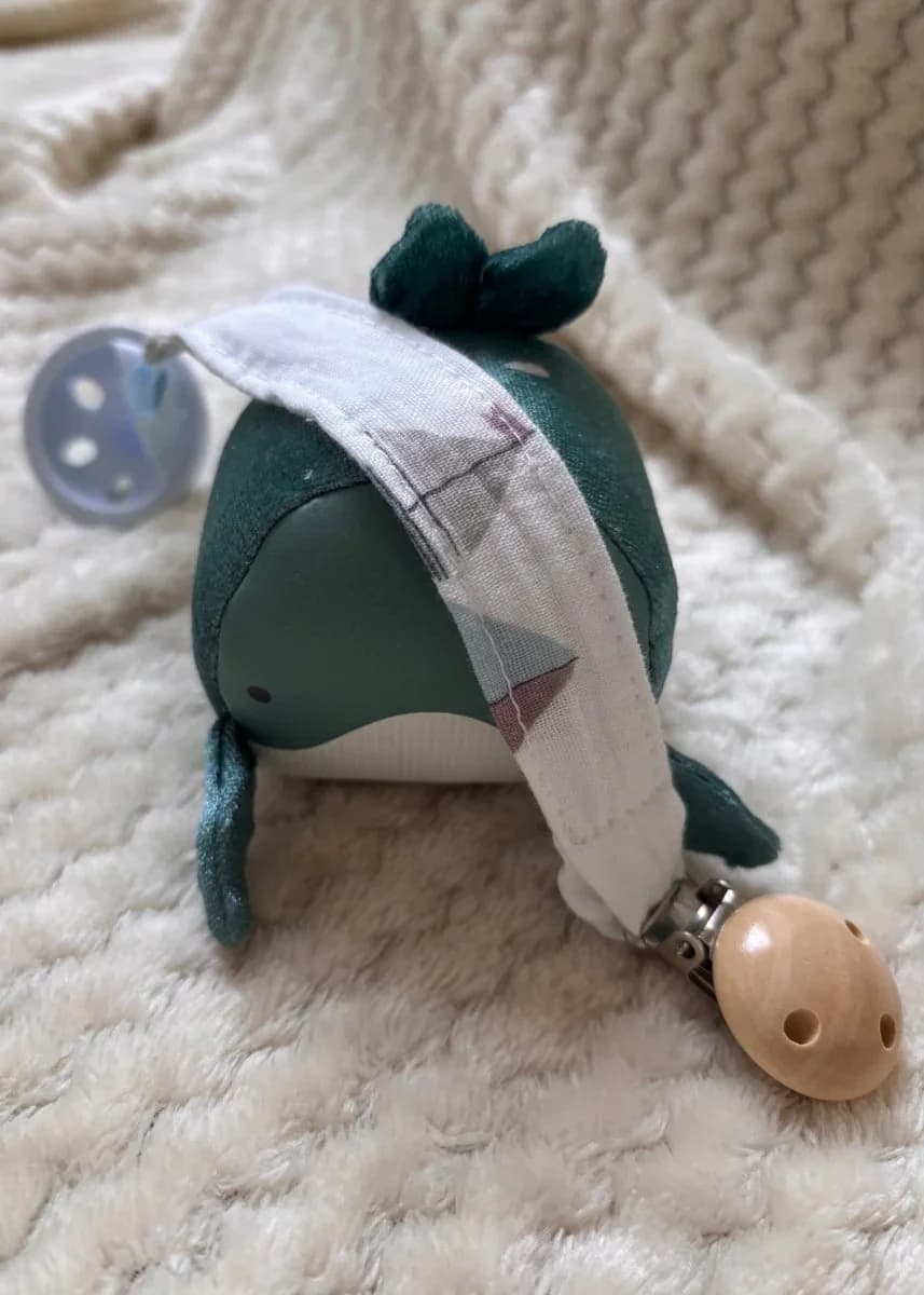 Attache-tétine en tissu à motifs discrets avec clip rond en bois clair, posée sur une peluche baleine.