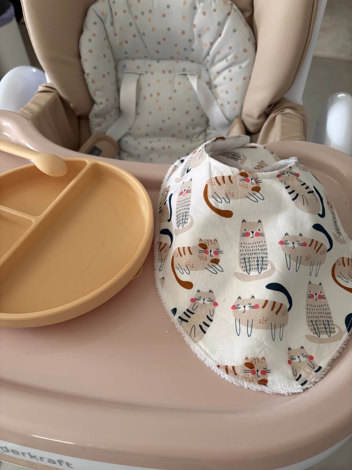 Bavoir bandana aux motifs de chats fermé en col, posé près d'une assiette sur une tablette de chaise haute.