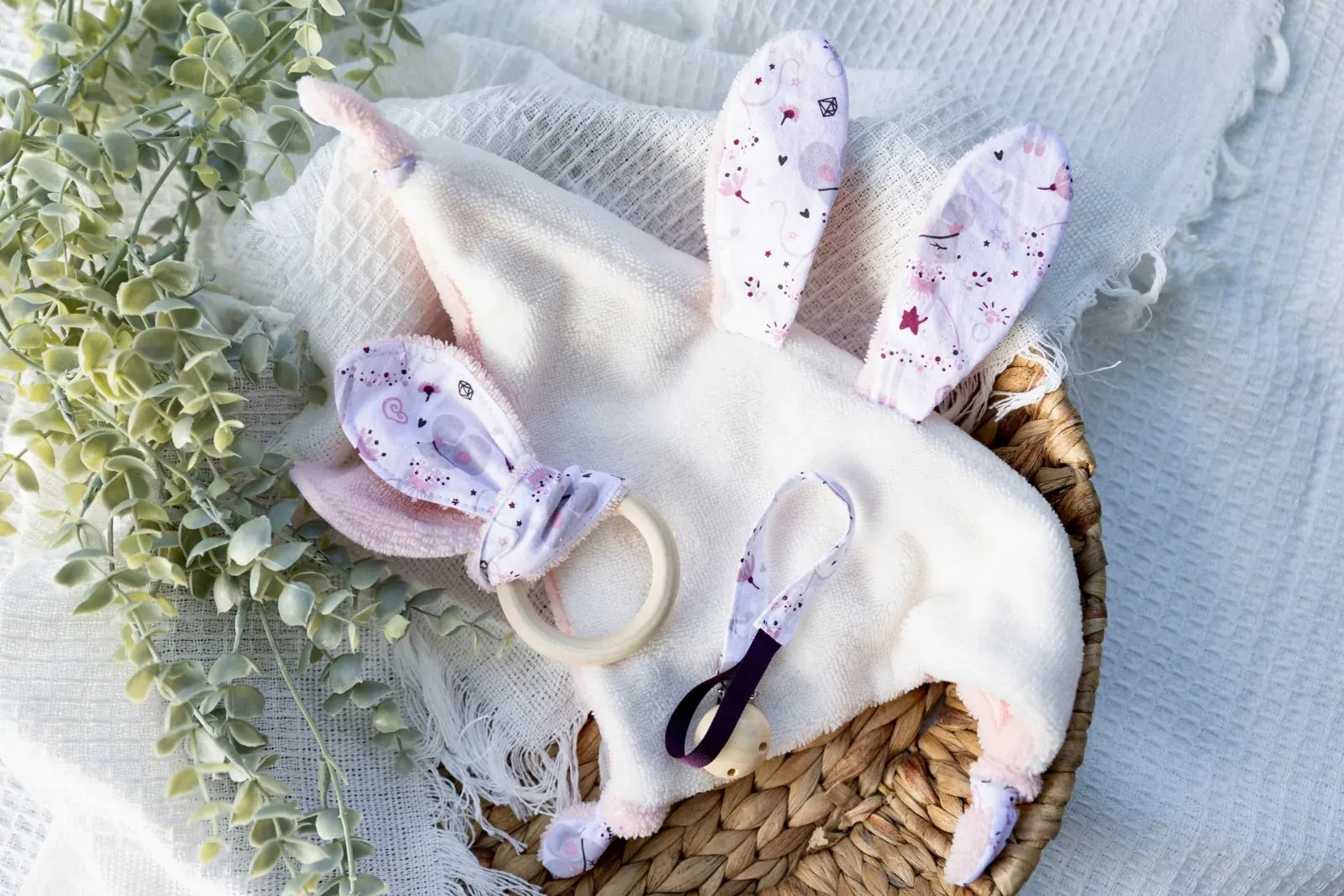 Doudou plat blanc et rose, anneau de dentition et attache-tétine à motif souris et étoiles dans un panier.