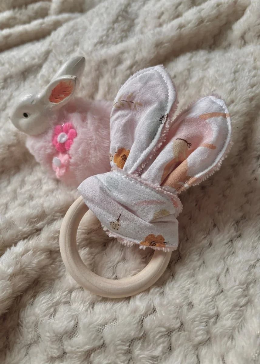 Anneau de dentition en bois avec oreilles en tissu fleuri doux, posé près d'un petit lapin décoratif.