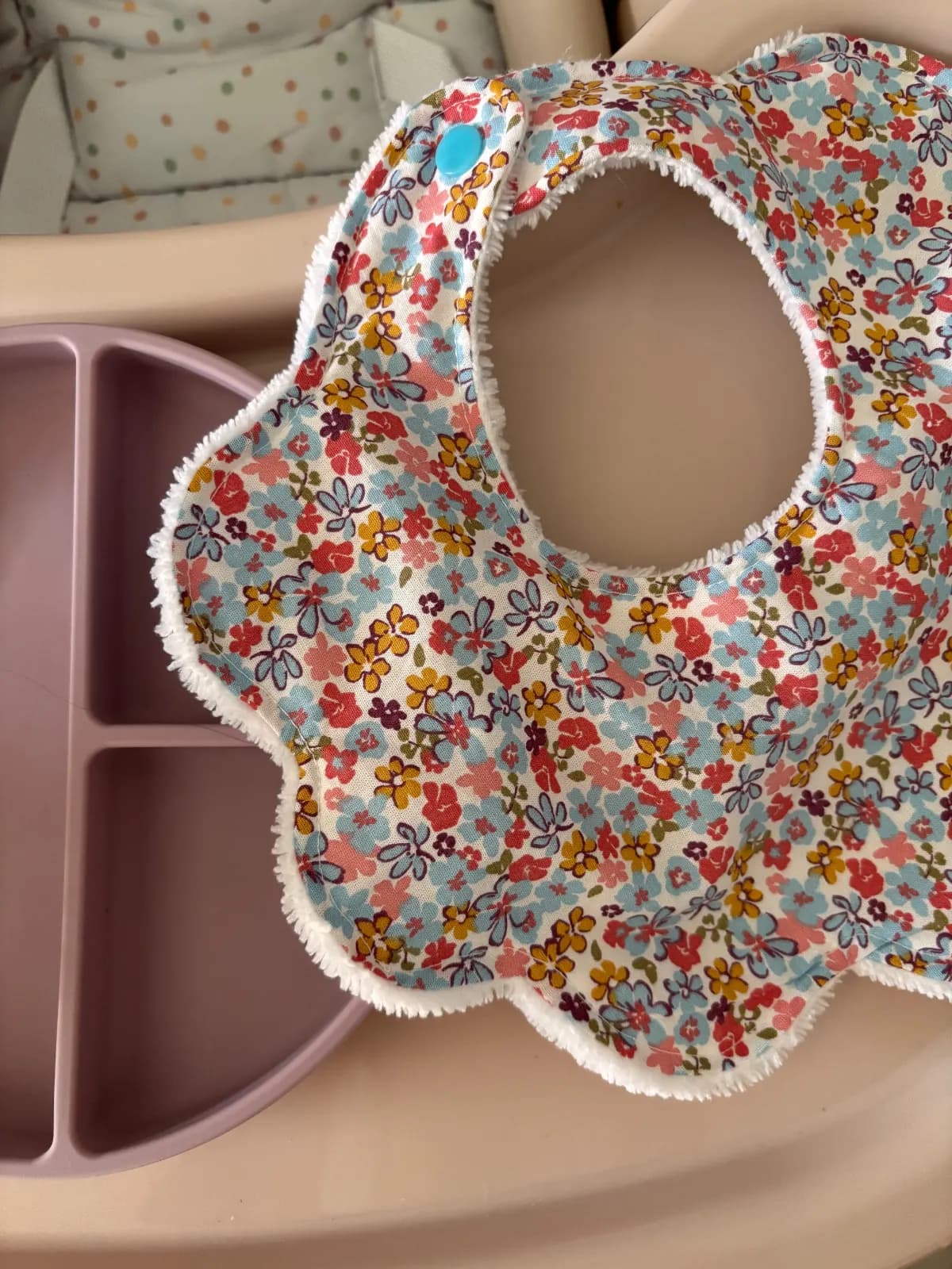 Gros plan sur le bavoir fleuri aux découpes de pétales et son bouton-pression turquoise