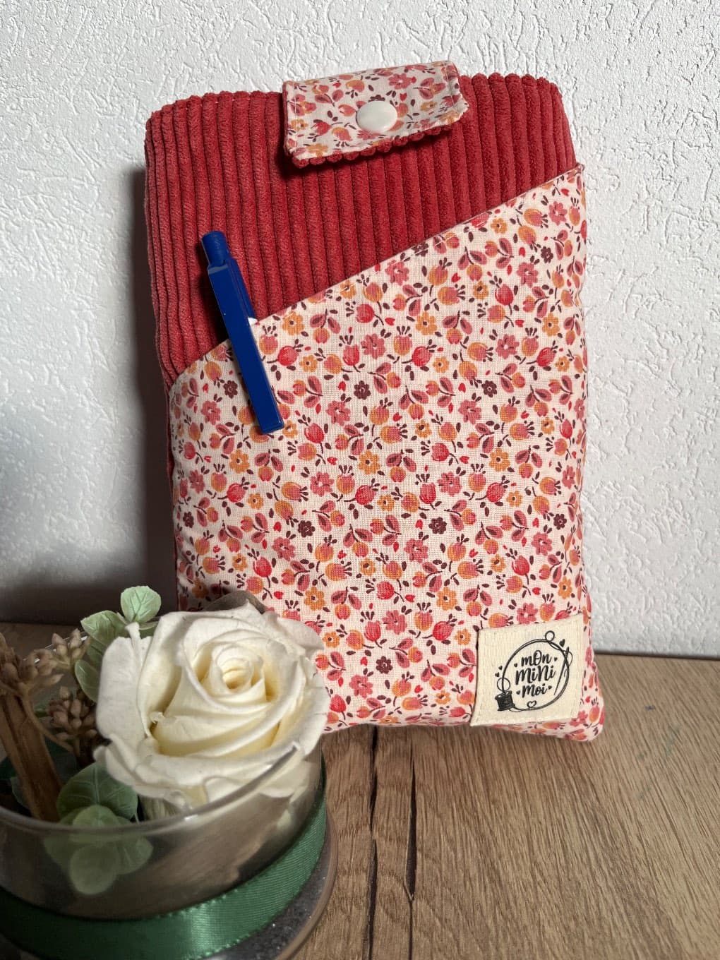 Pochette de livre debout avec stylo bleu glissé dans la poche avant en tissu fleuri.