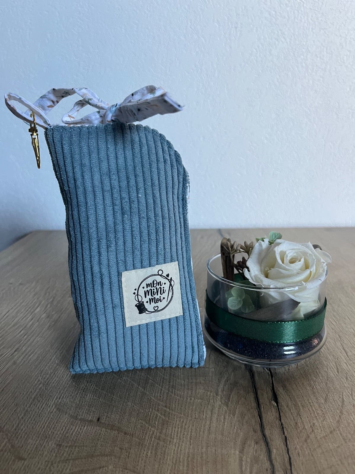 Étui à lunettes vertical en velours côtelé bleu avec nœud fleuri, posé près d'une rose blanche.