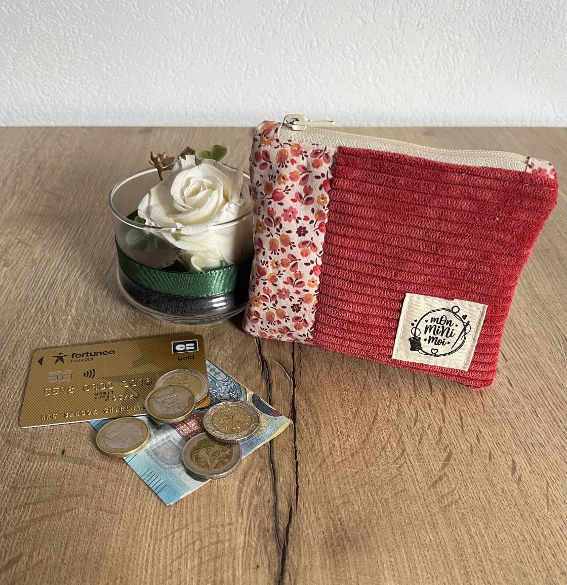 Porte-monnaie rose framboise et tissu fleuri posé près de pièces, d'une carte et d'un vase avec rose