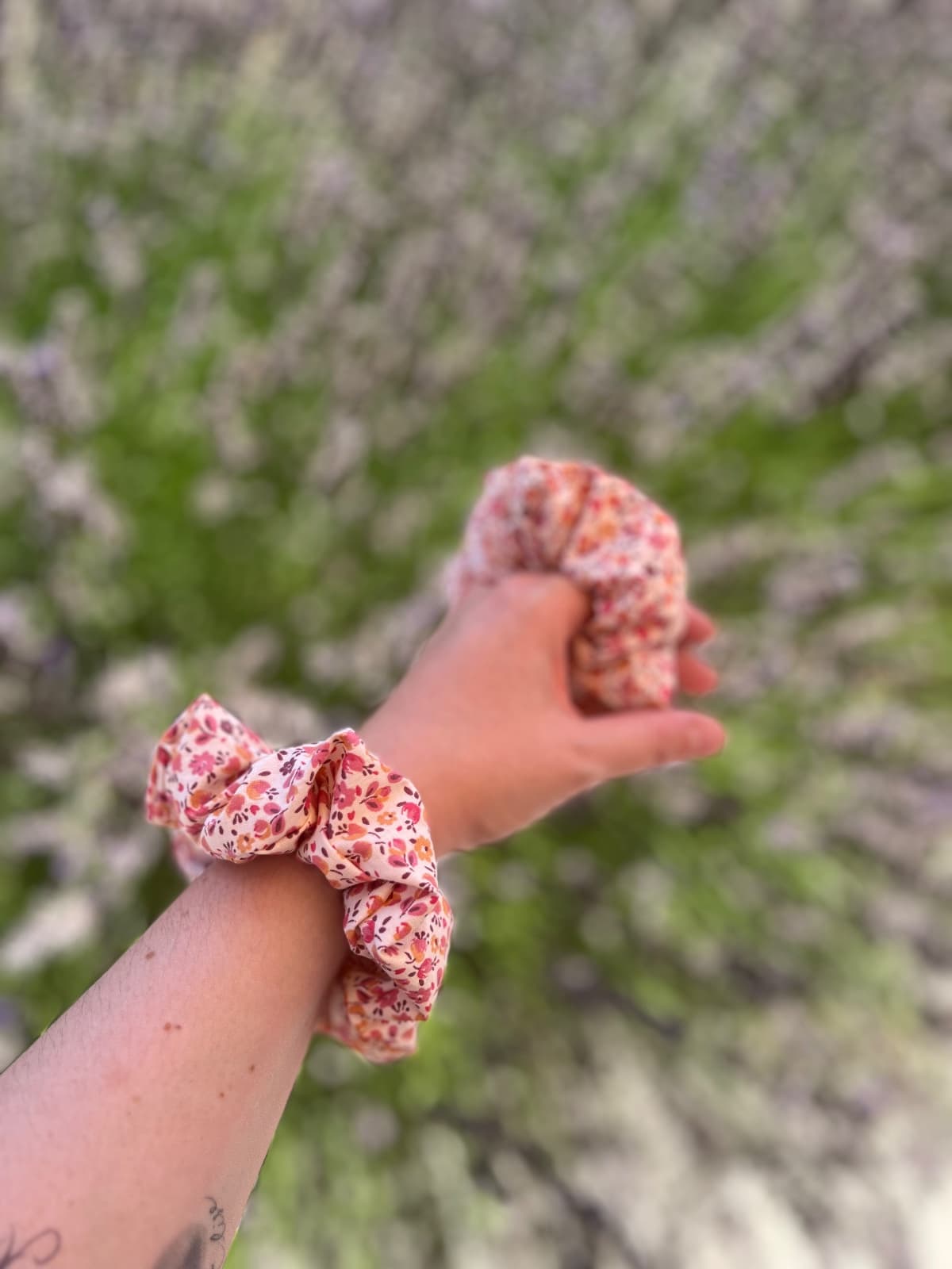 Chouchou à motif fleuri rose porté au poignet en extérieur sur fond de végétation floue.