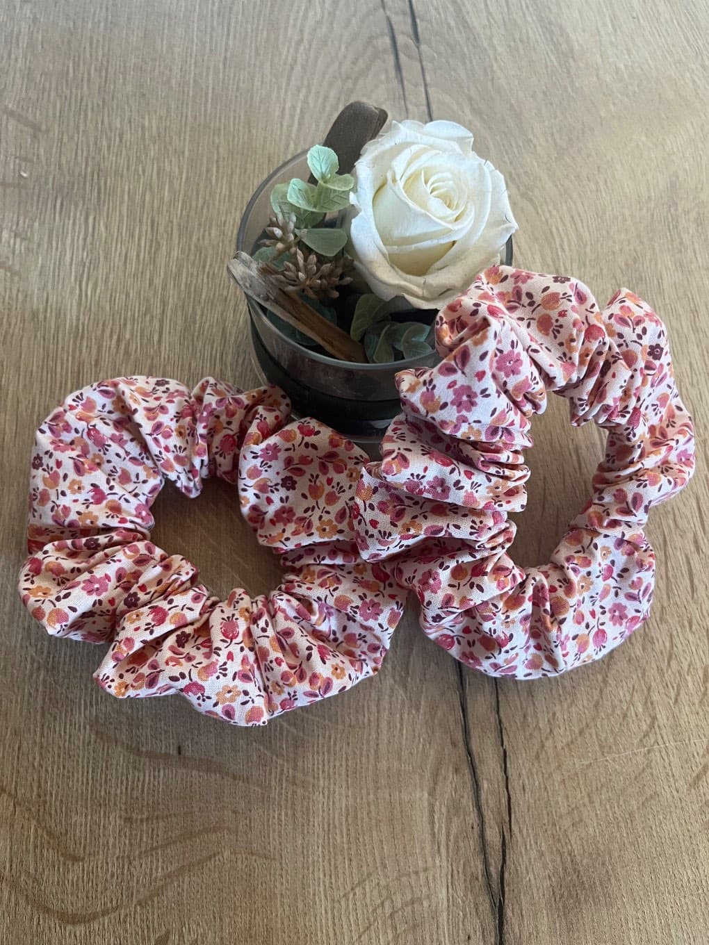 Deux chouchous froncés à motif floral rose et bordeaux posés sur du bois près d'une rose blanche décorative.