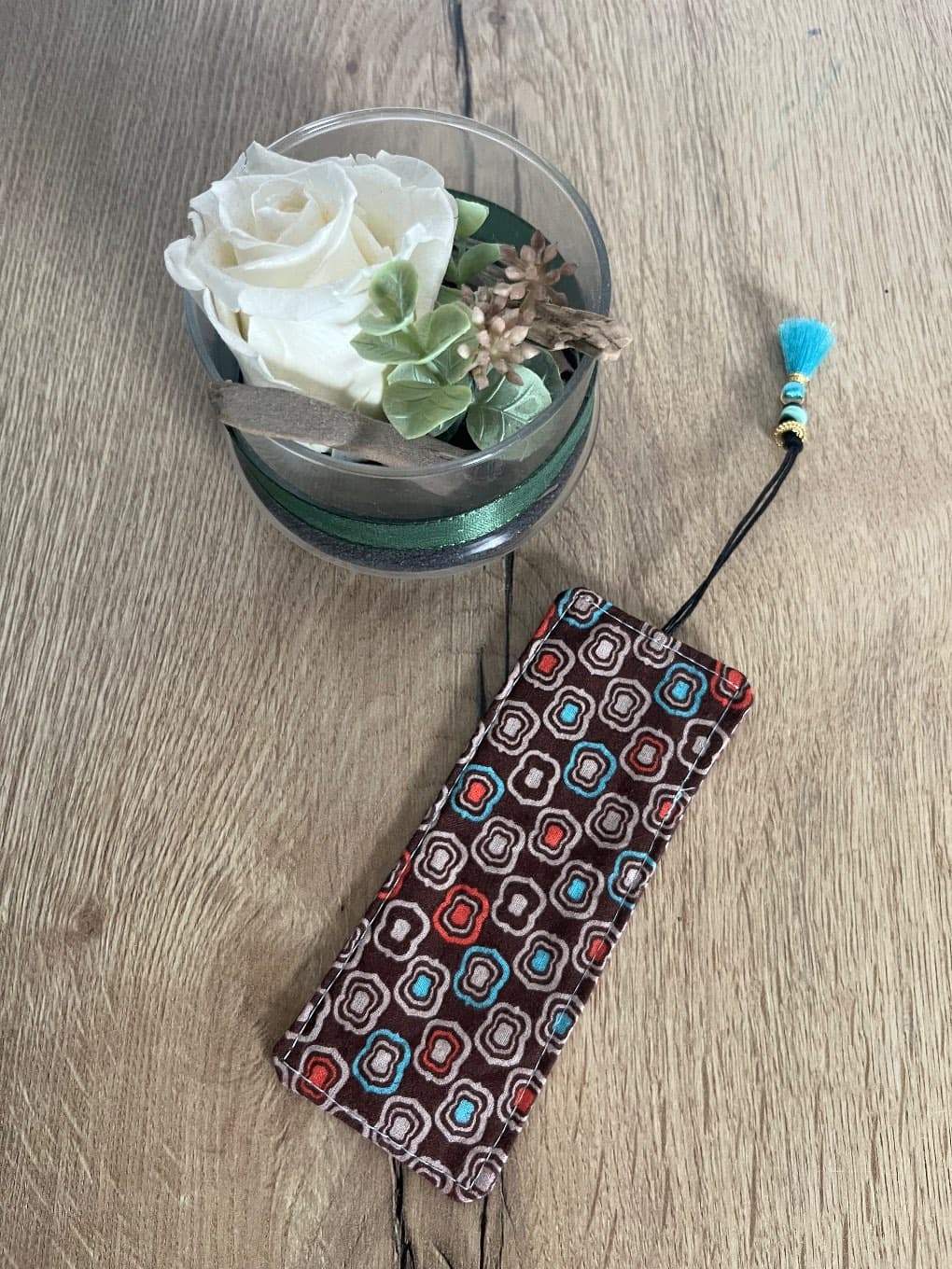Marque-page cousu rectangulaire à motifs rétro marron et pompon turquoise posé à plat sur du bois
