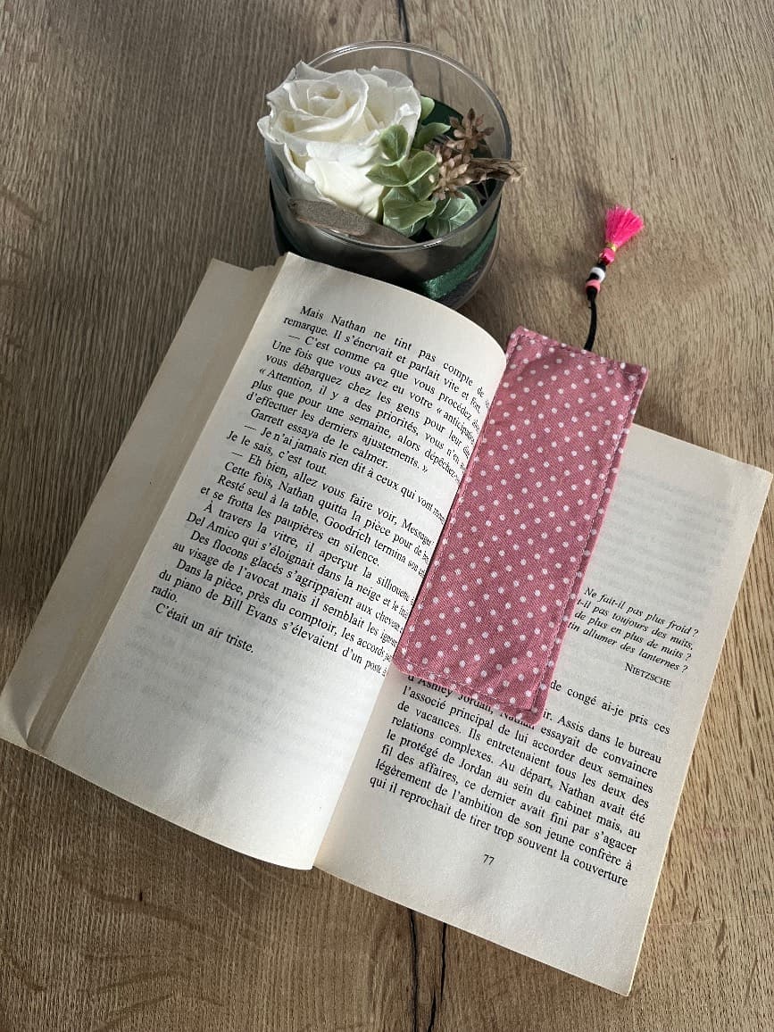 Marque-page rectangulaire en tissu rose à pois blancs avec cordon et pompon, posé sur une table en bois.